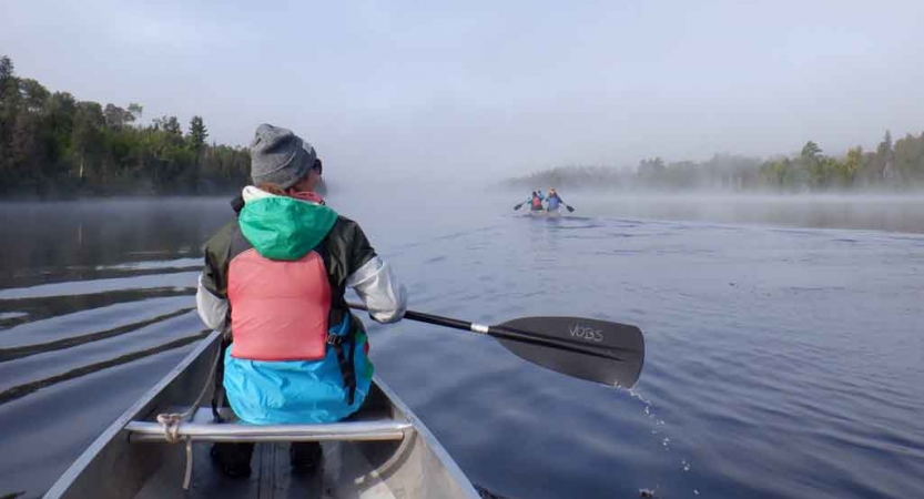 a young person sits in the front of a canoe holding a paddle. The water is calm and there is a canoe distorted by fog in the distance. 